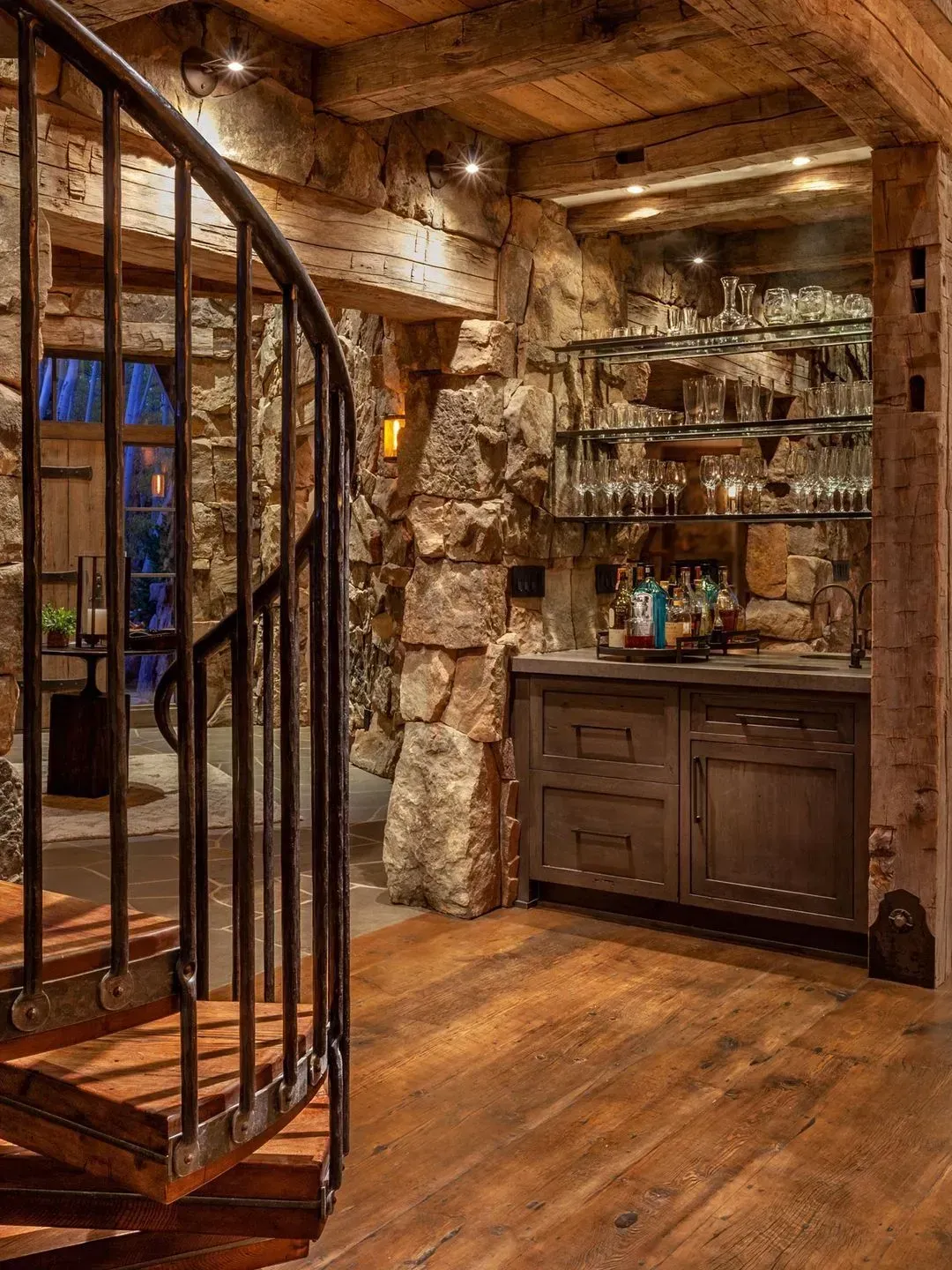 Rustic bar area with stone walls, wooden floors, spiral staircase. Shelves with glasses, bottles.
