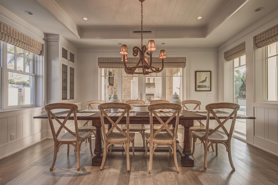 Dining room with a long wooden table, chairs, chandelier, and windows with shades.