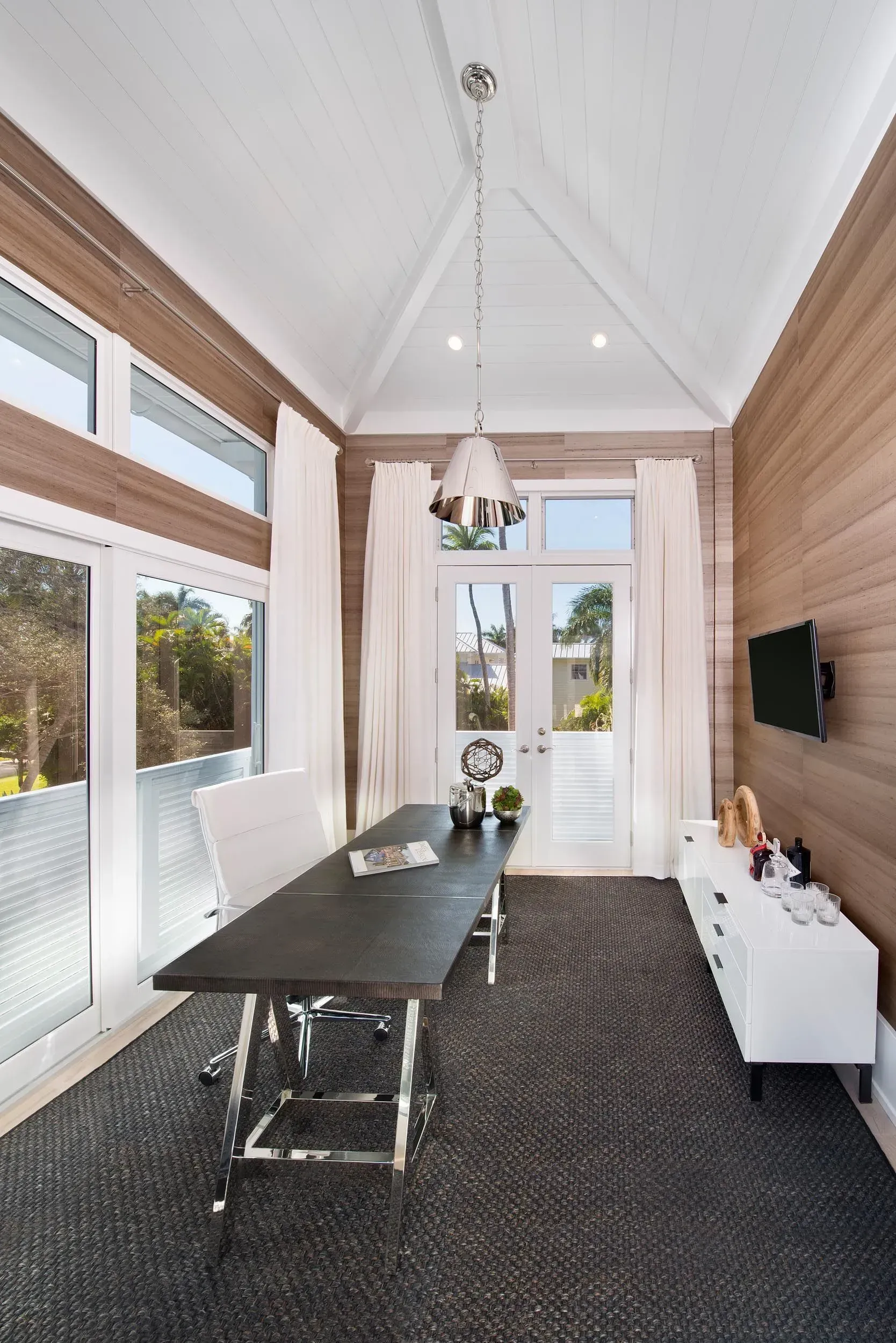 Office with wood paneling, large windows, dark rug, desk, white storage, and a pendant light.