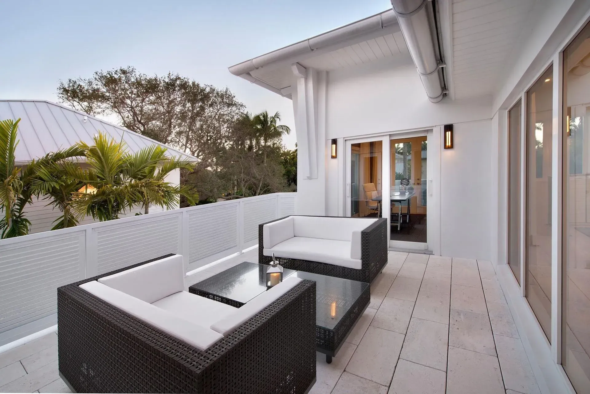 Outdoor patio with white furniture and glass-topped table, overlooking a white-roofed house and trees.
