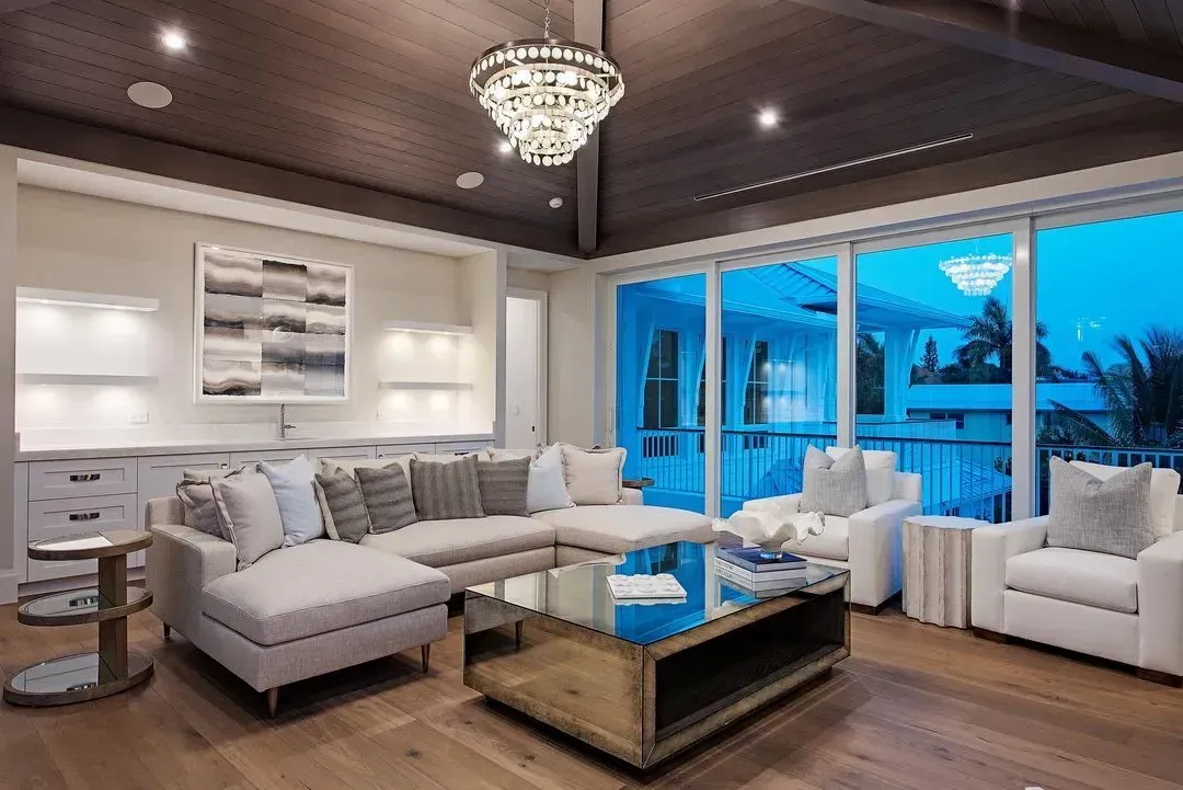 Living room with white furniture, wooden floors, large windows, and a chandelier.