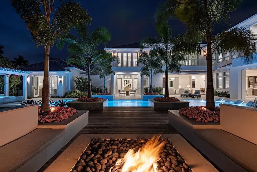 Night view of a luxury home with a pool, fire pit, and palm trees; white exterior lit with warm lights.