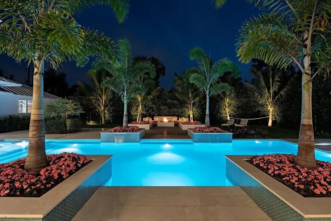 Nighttime view of a turquoise pool with palm trees, flowerbeds, and uplighting.