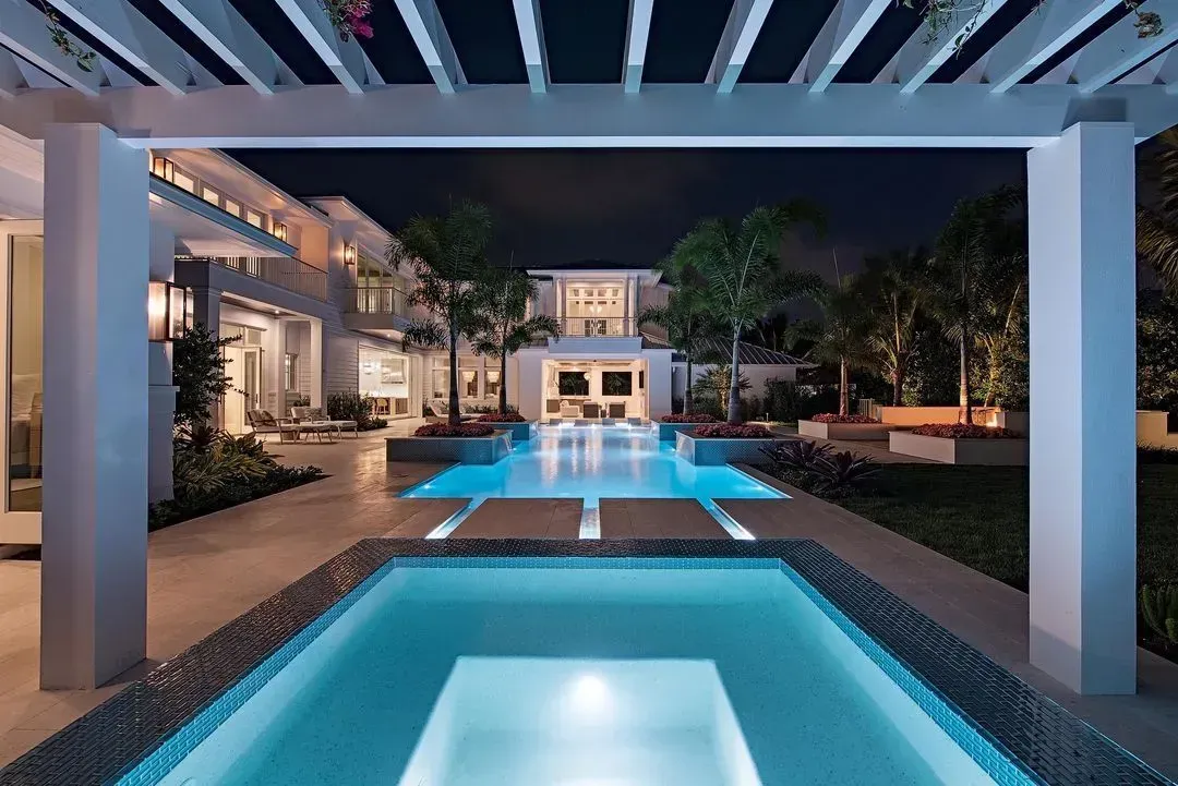 Night view of a luxury home with lit pool, spa, and pergola.