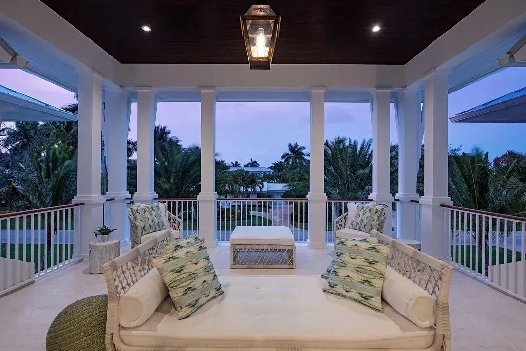 Balcony with seating, white columns, railing, and a view of trees at dusk.