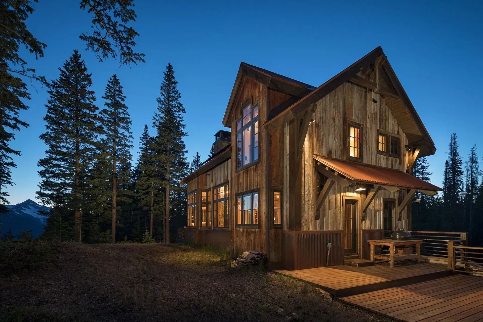 Rustic wooden cabin at dusk, illuminated windows, nestled in a forest with mountain backdrop.