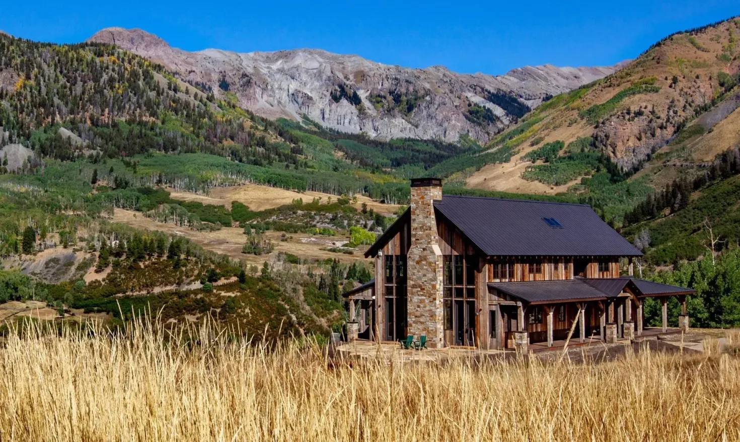 Rustic cabin nestled in a valley with mountains in the background under a blue sky, surrounded by golden grass.