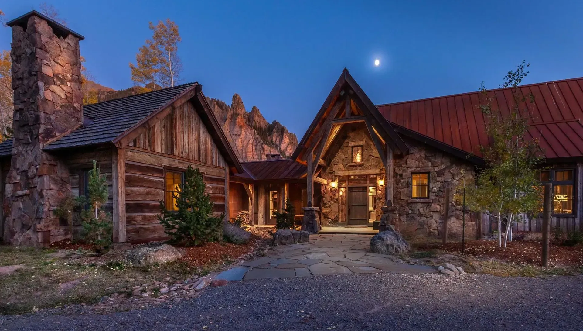 Rustic stone and wood lodge at dusk, lit interior with chimney and moon visible.
