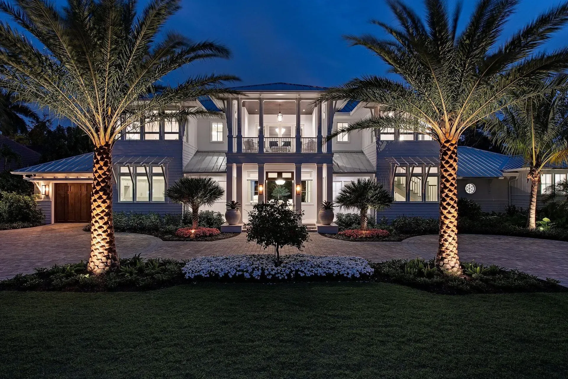 Lit-up luxury home at night, flanked by palm trees, and lit landscaping. Gravel driveway and manicured lawn.