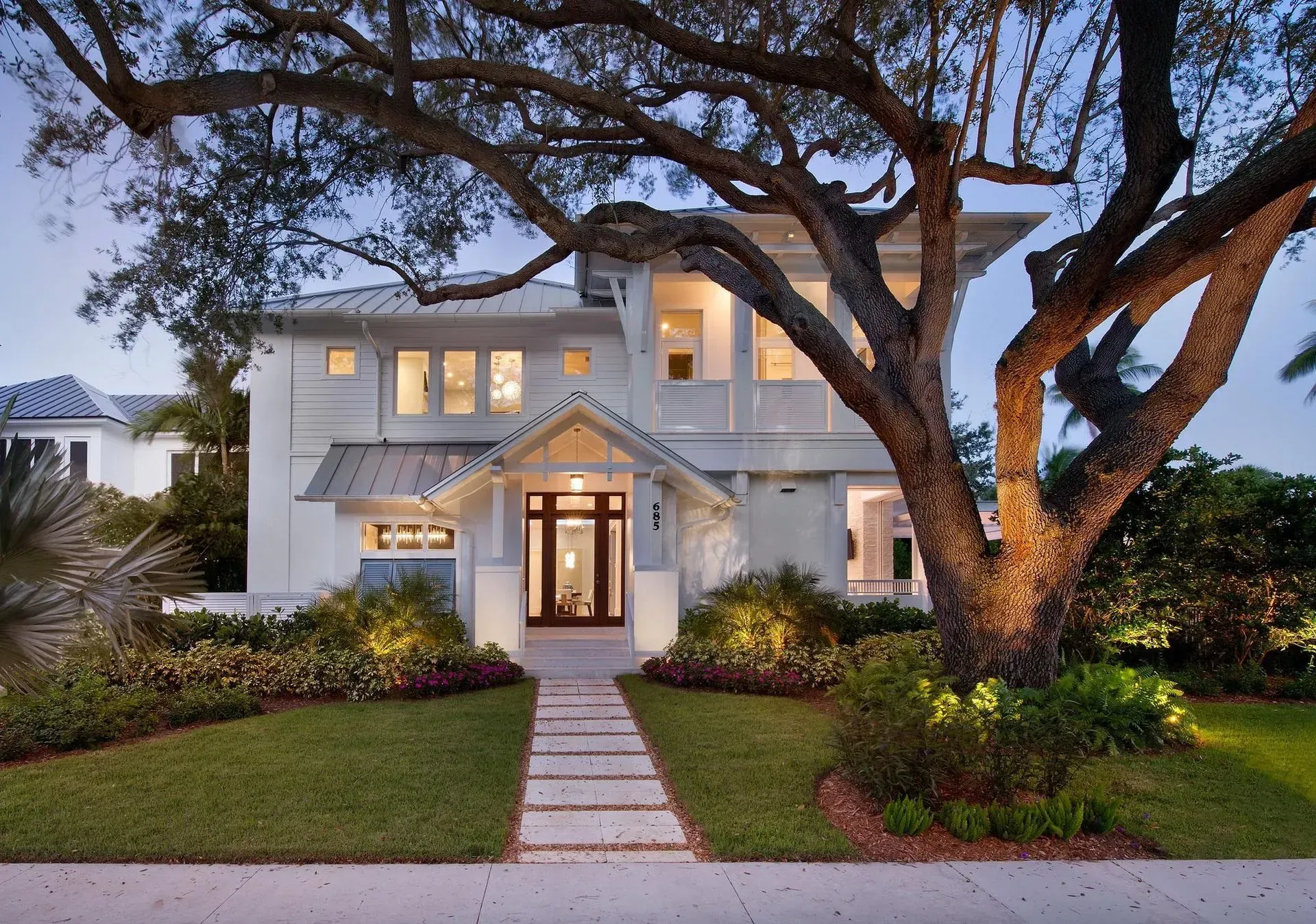 White two-story house with a lit pathway, large tree, and well-maintained landscaping at dusk.