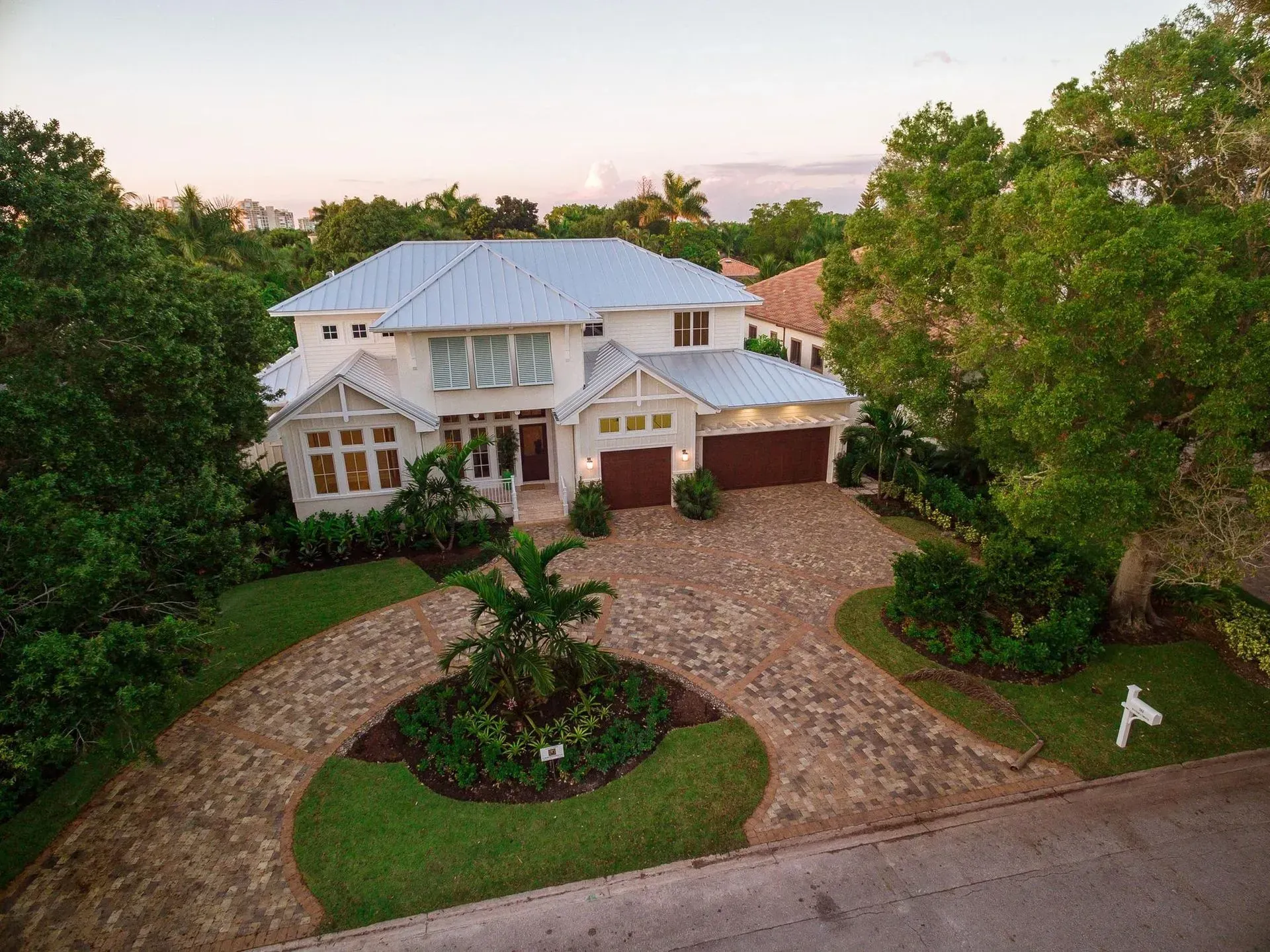Two-story white house with light blue roof and brown garage doors surrounded by trees and a circular driveway.