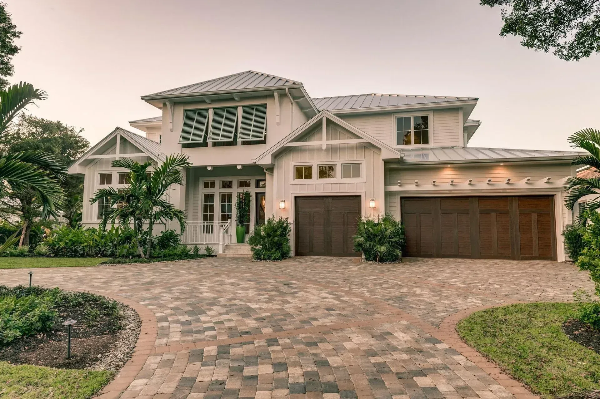 White two-story house with dark wood garage doors, brick driveway, and lush greenery.