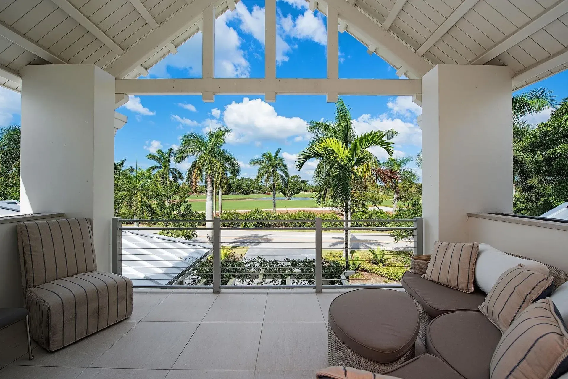 Balcony overlooking a golf course, furnished with seating and pillows, with a blue sky and palm trees.