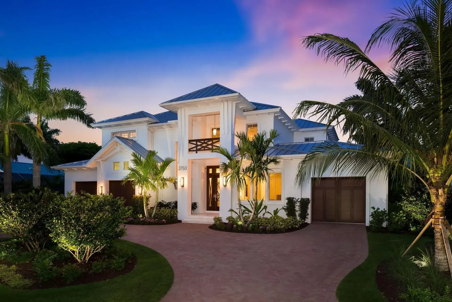 White luxury home with a circular driveway and palm trees at dusk.