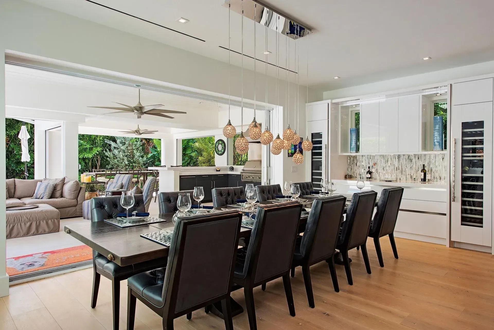 Elegant dining room with long table, chairs, and hanging lights, open to living area and outdoors.