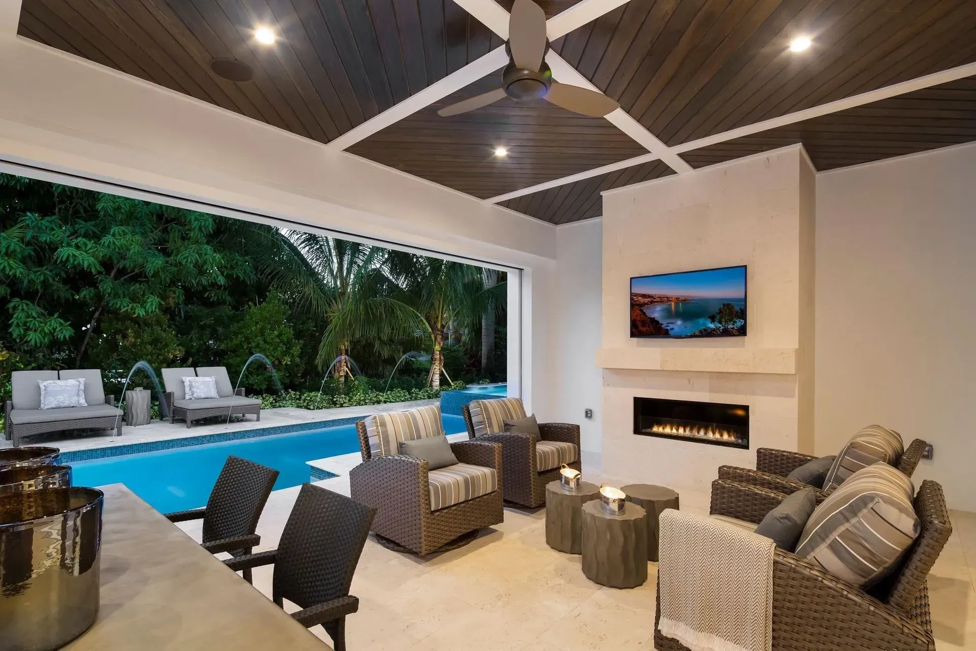 Outdoor living area with pool, fireplace, TV, and seating under a wood-paneled ceiling.