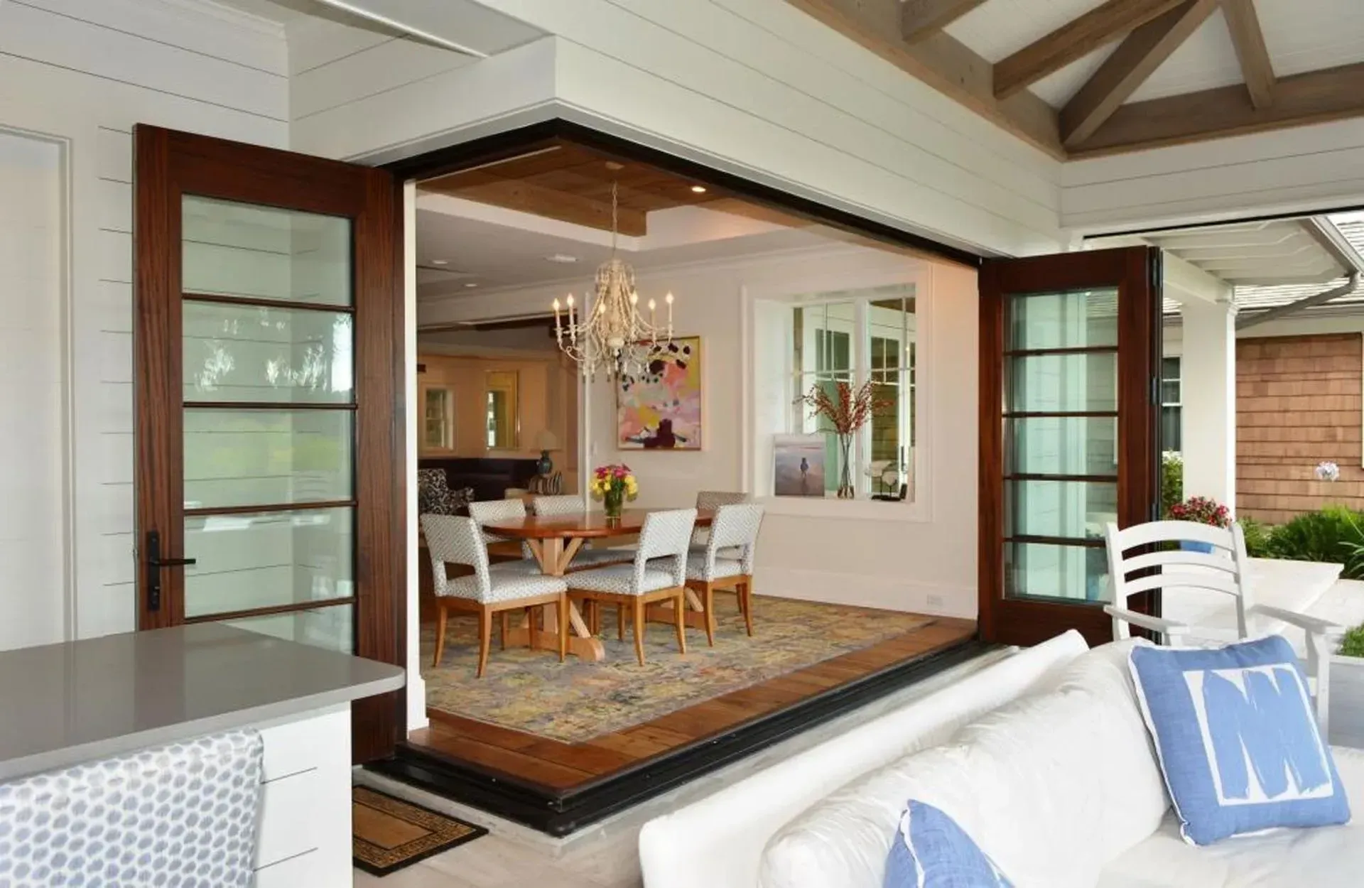 Open indoor/outdoor dining room with dark wood doors, table, chairs, chandelier, and a white sofa.