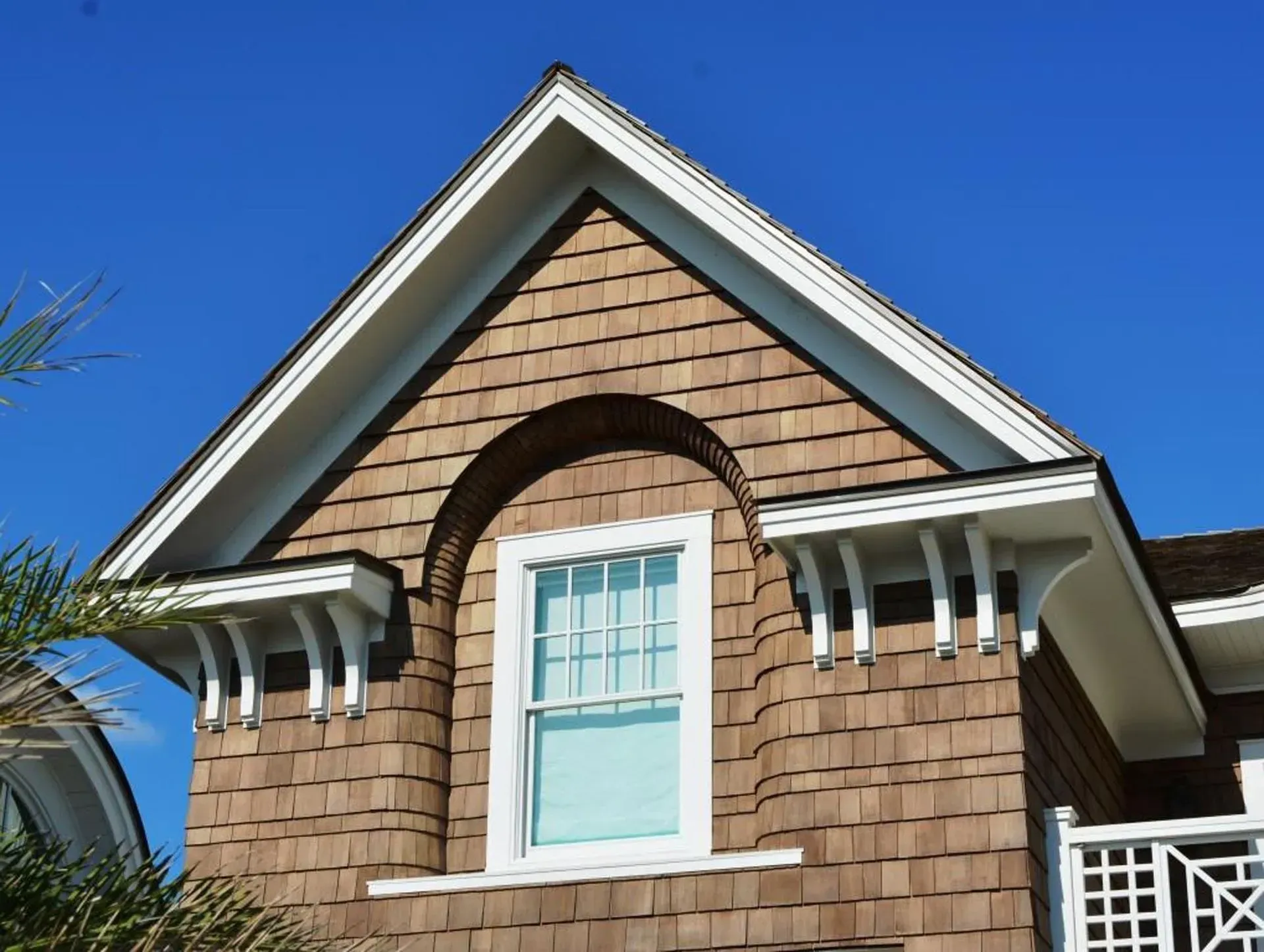 Triangular brown shingled house gable with white trim and window against blue sky.