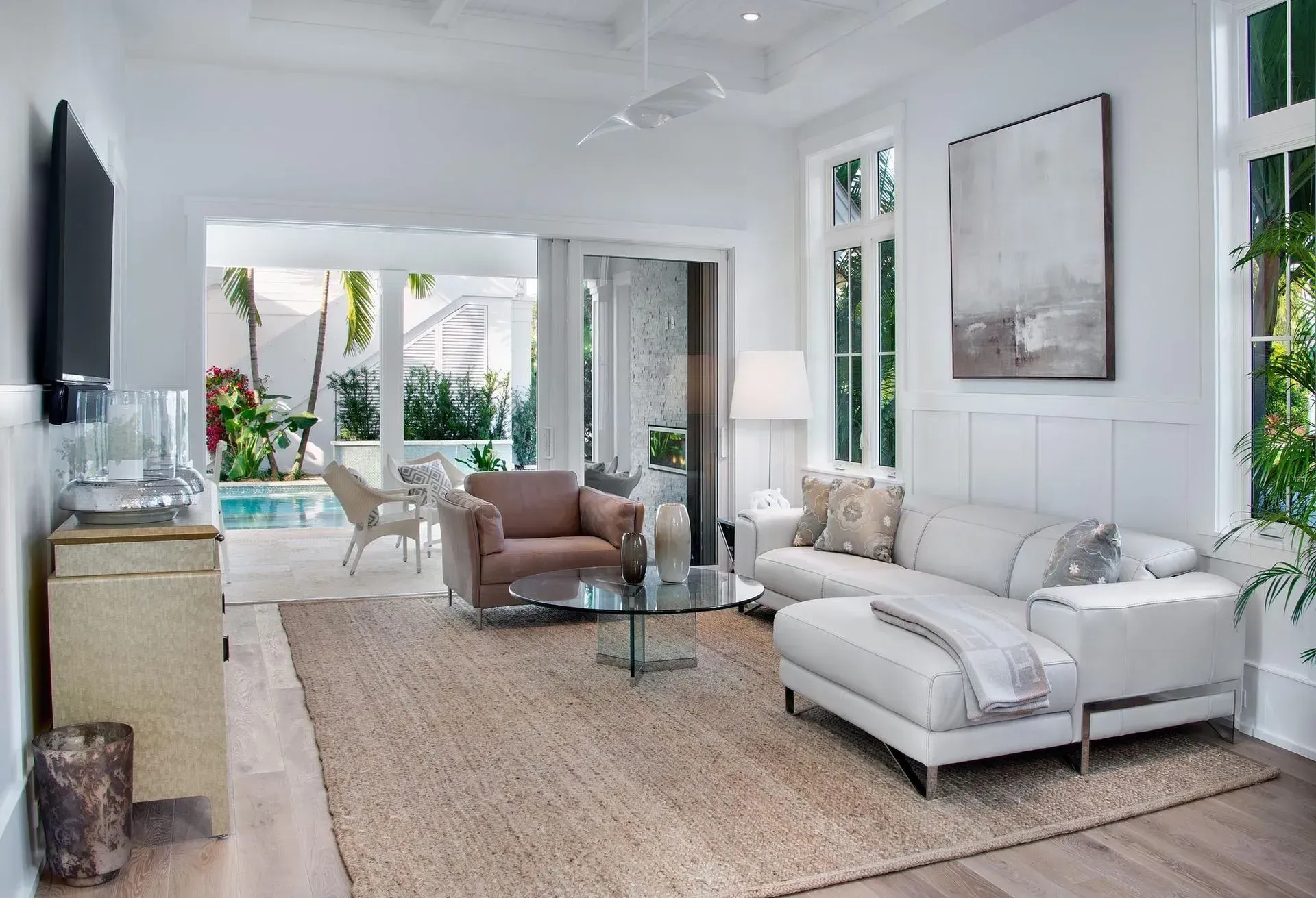 Bright white living room with pool view, sofa, armchair, jute rug, glass coffee table, and large abstract artwork.