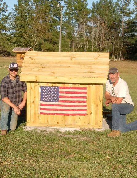 Two men are kneeling in front of a small wooden house with an american flag on it.