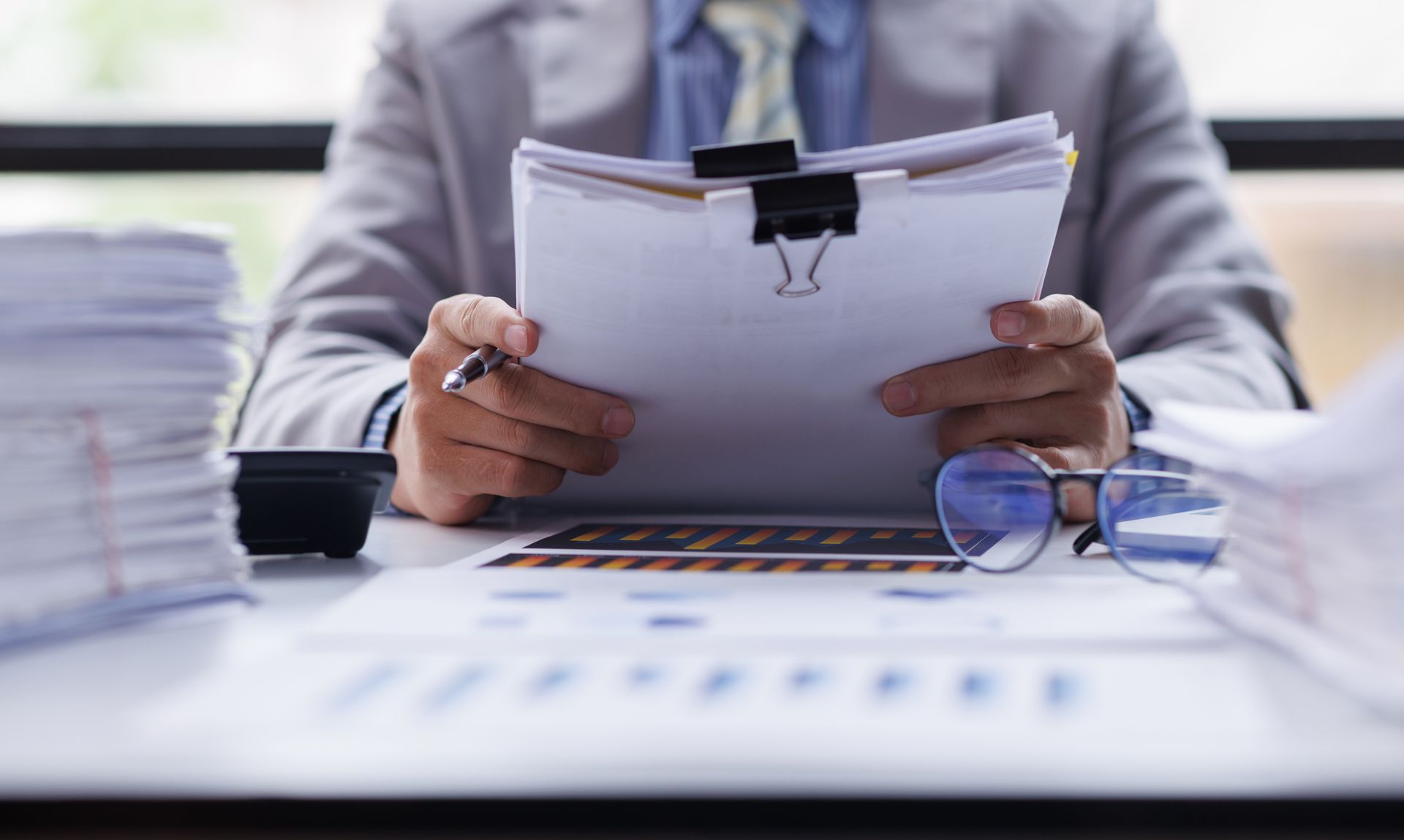 Person reviewing paperwork at a desk with a clipboard, stacks of papers, and glasses nearby