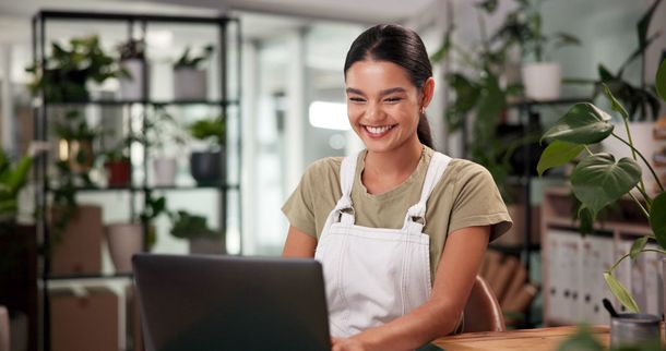 Smiling person in an apron using a laptop in a bright plant-filled shop