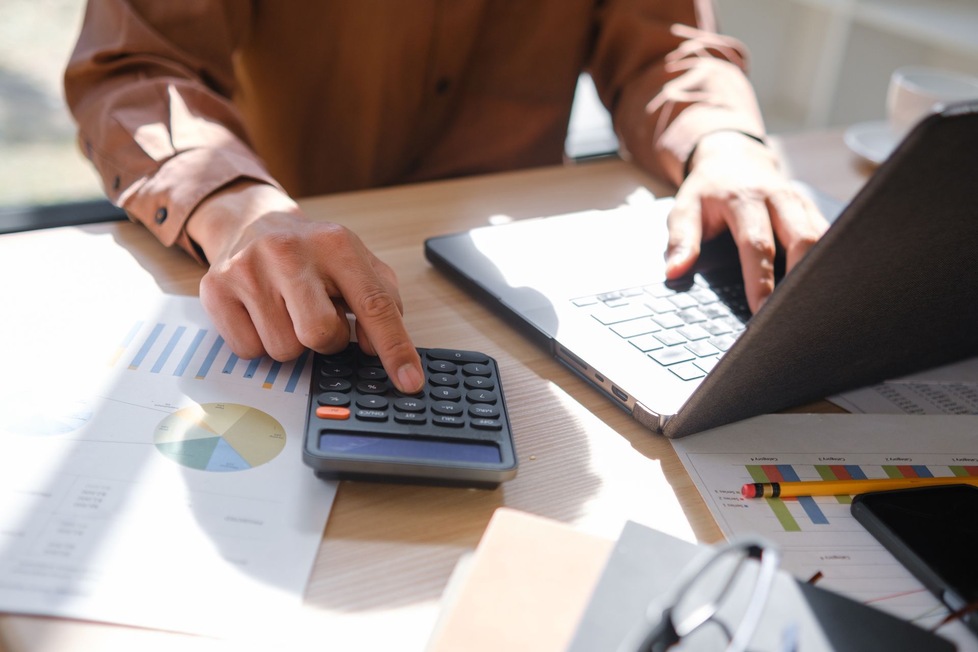 Hands using a calculator beside a laptop on a desk with paperwork and charts