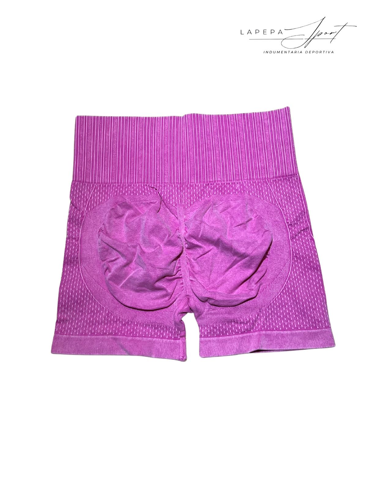 Mujer con pantalones cortos rosas y sujetador deportivo negro, posando frente a una pared de ladrillos blancos.