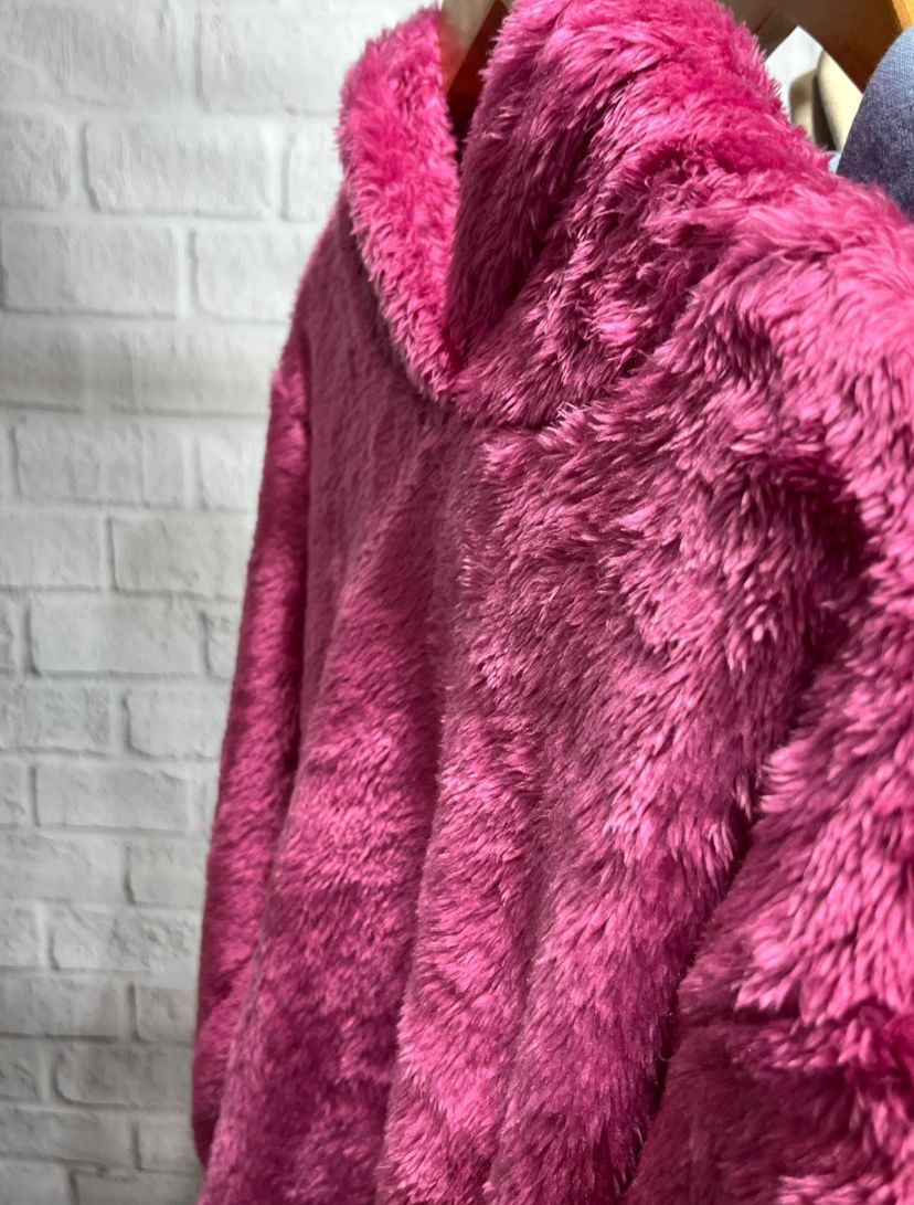 Mujer con una sudadera con capucha rosa afelpada, con las manos en el bolsillo delantero, de pie frente a un fondo blanco.