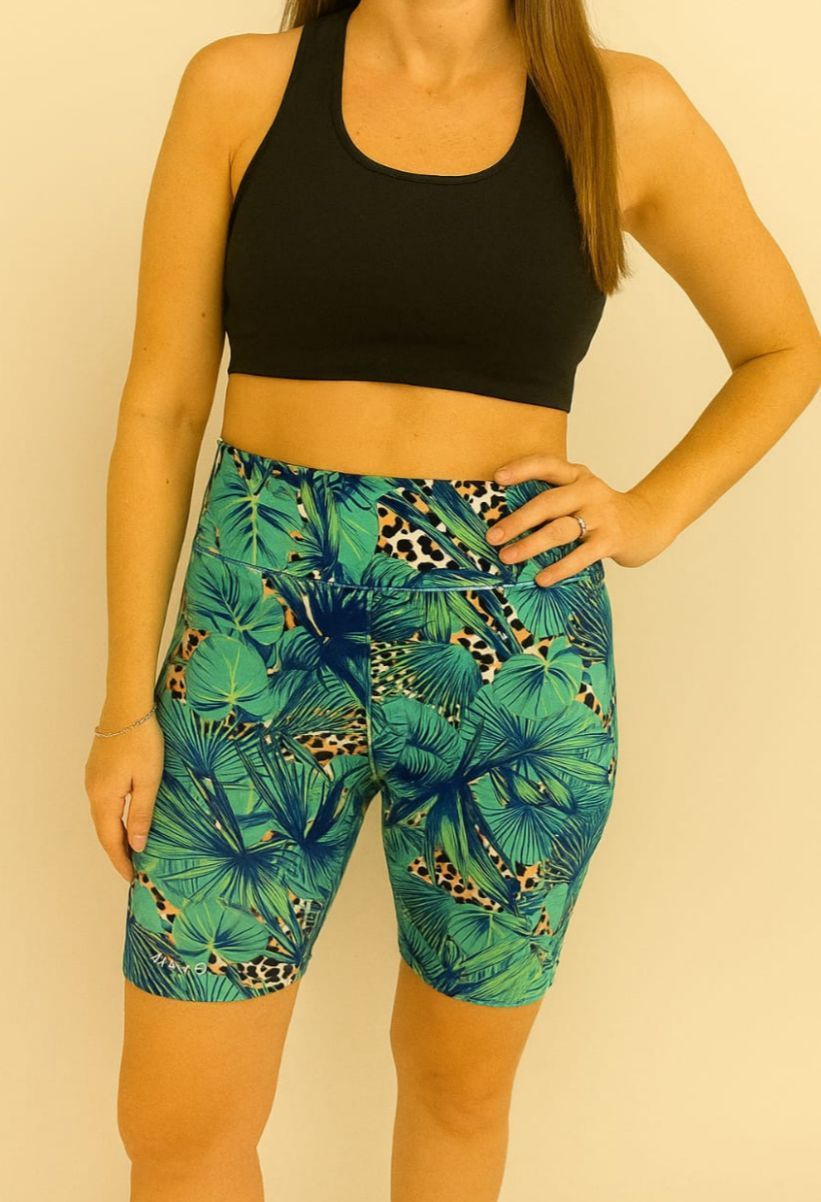Mujer con sujetador deportivo negro y pantalones cortos de ciclista estampados en verde, con la mano en la cadera.