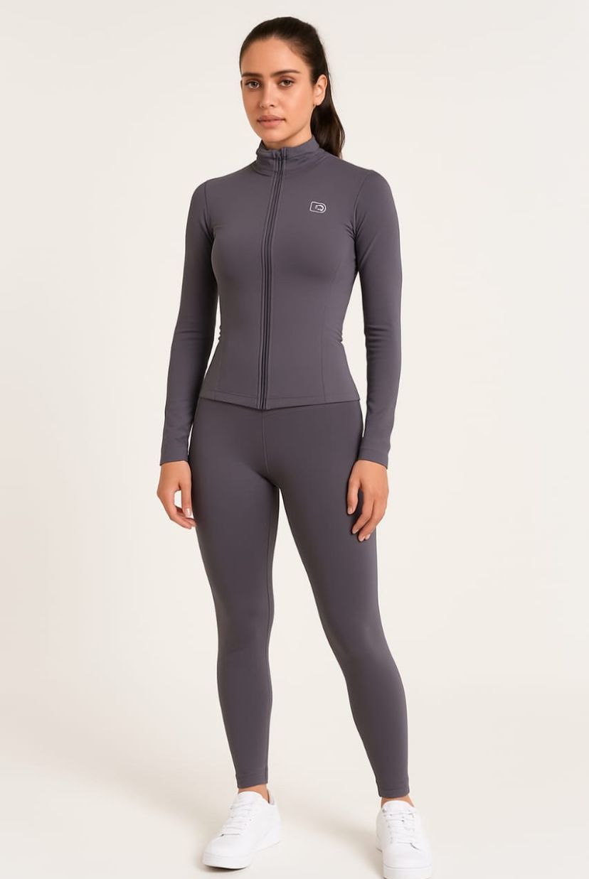 Mujer vestida con ropa deportiva gris, de pie, con una chaqueta con cremallera y mallas, con zapatillas blancas.