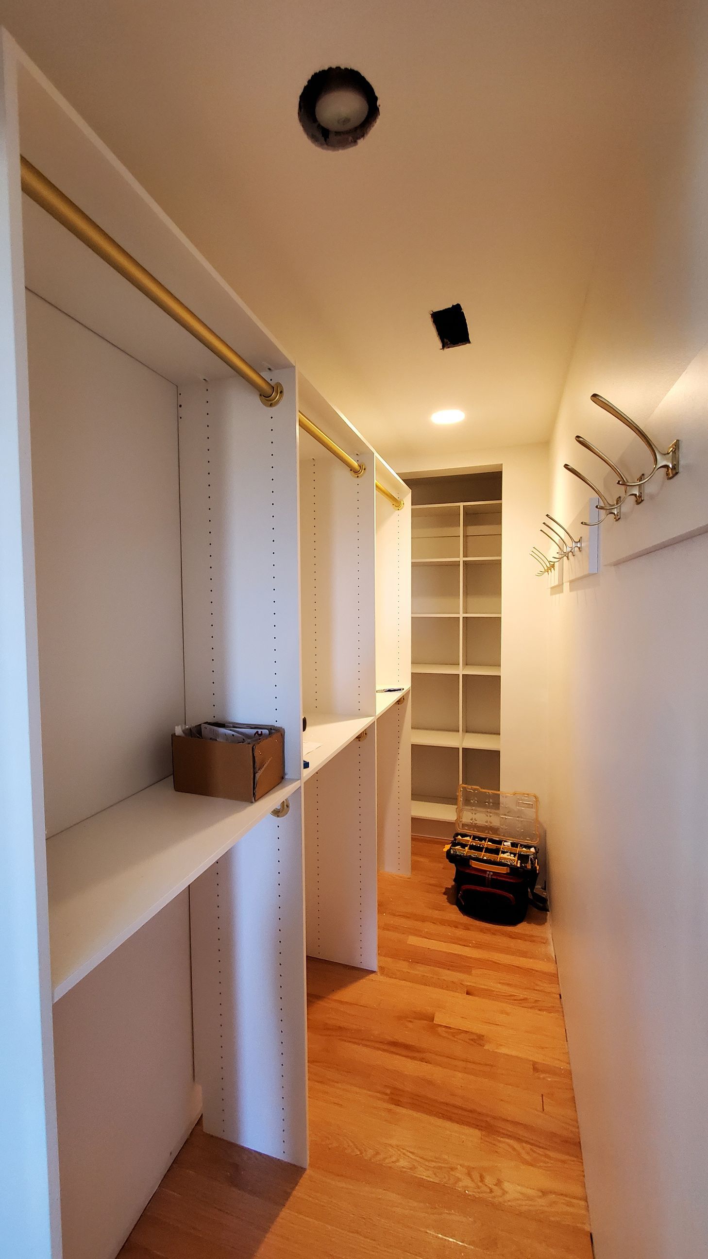A narrow, unfinished walk-in closet with white wooden shelving units, gold clothing rods, and wall-mounted hooks.