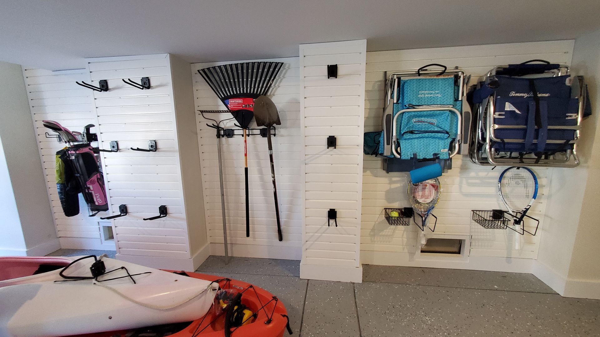 Garage wall with white slatwall storage holding tools, beach chairs, and a backpack, with a kayak in the foreground.
