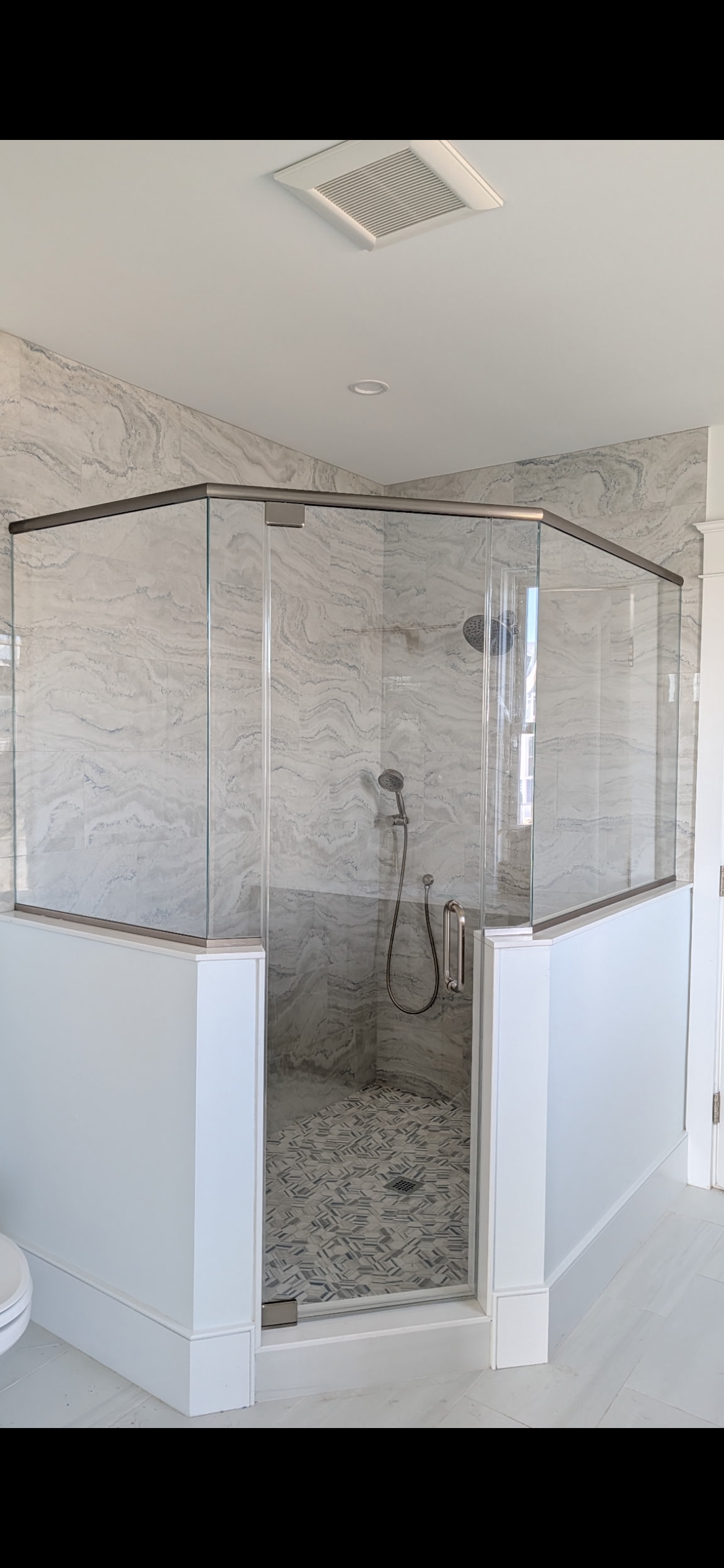 A walk-in shower with glass walls, tiled floors with pebble accents, and white pony walls in a bathroom.