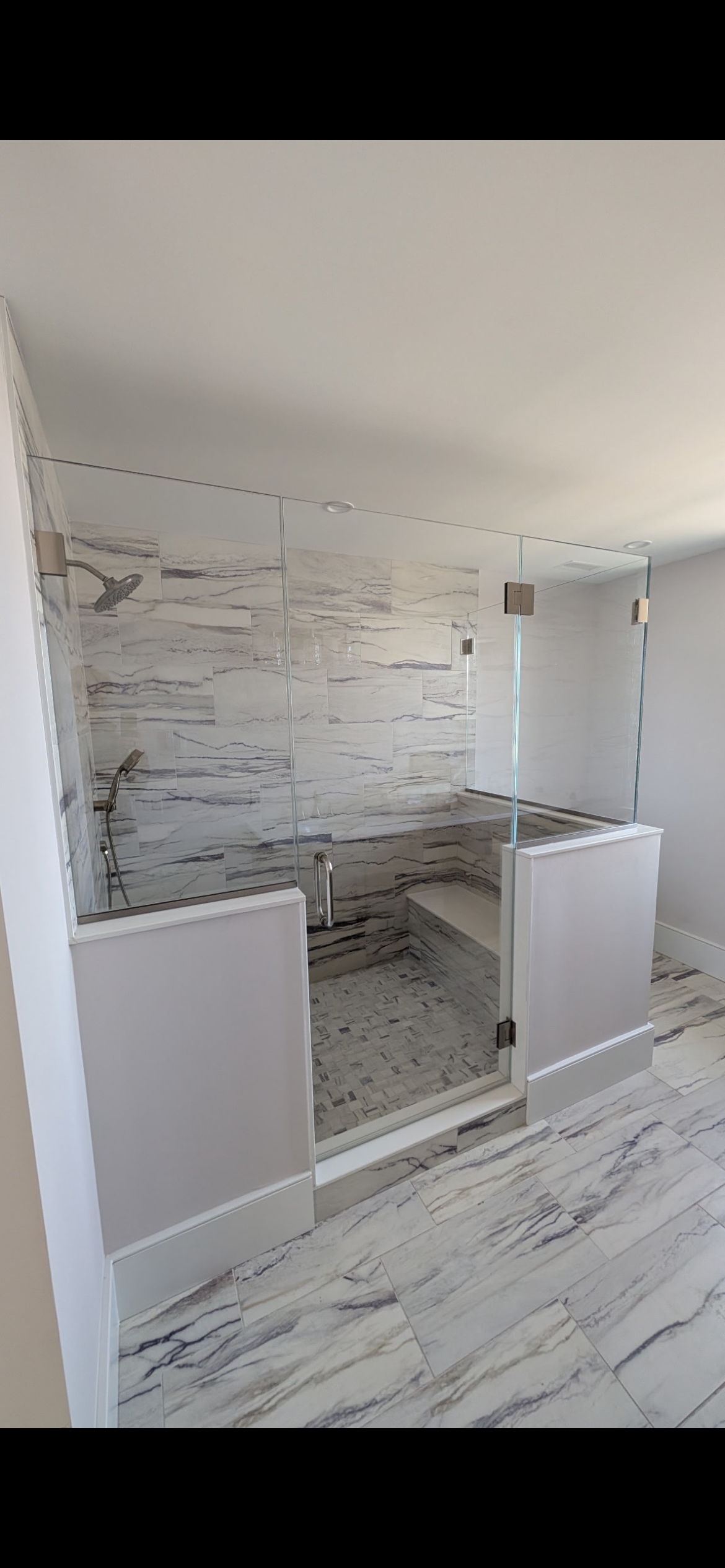 A modern, marble-tiled shower stall with glass walls, a bench, and a low partial wall in a bright bathroom.