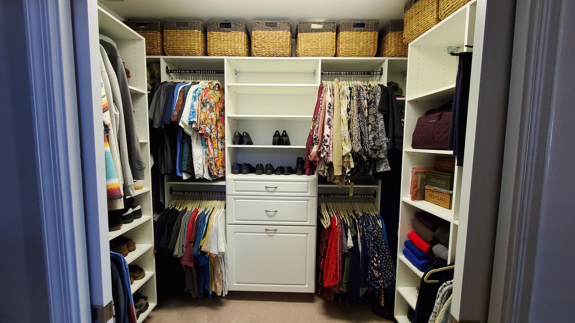 A white walk-in closet organized with hanging clothes, shelves, drawers, and woven storage bins on the top shelf.