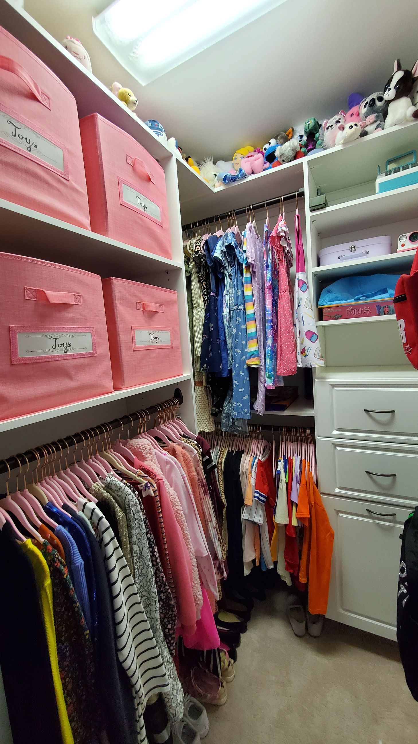 A walk-in closet with shelves holding pink storage bins, hanging clothes in various colors, and drawers on the right.