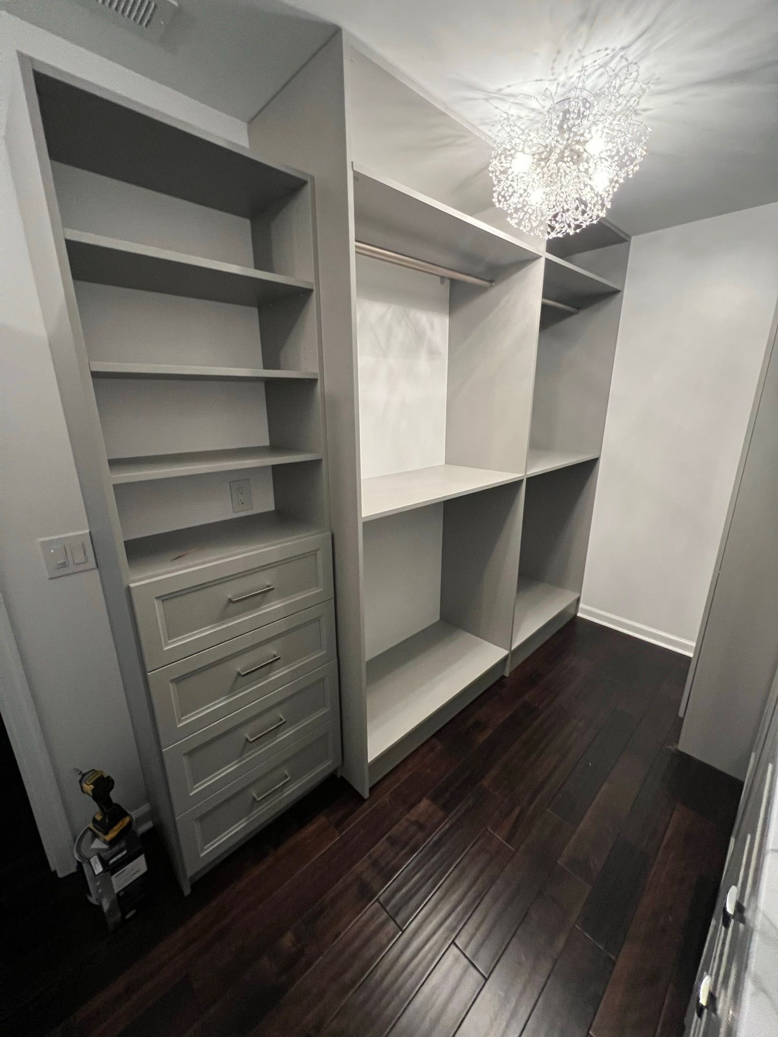 A walk-in closet featuring light gray shelving, drawers with silver pulls, dark hardwood floors, and a modern chandelier.