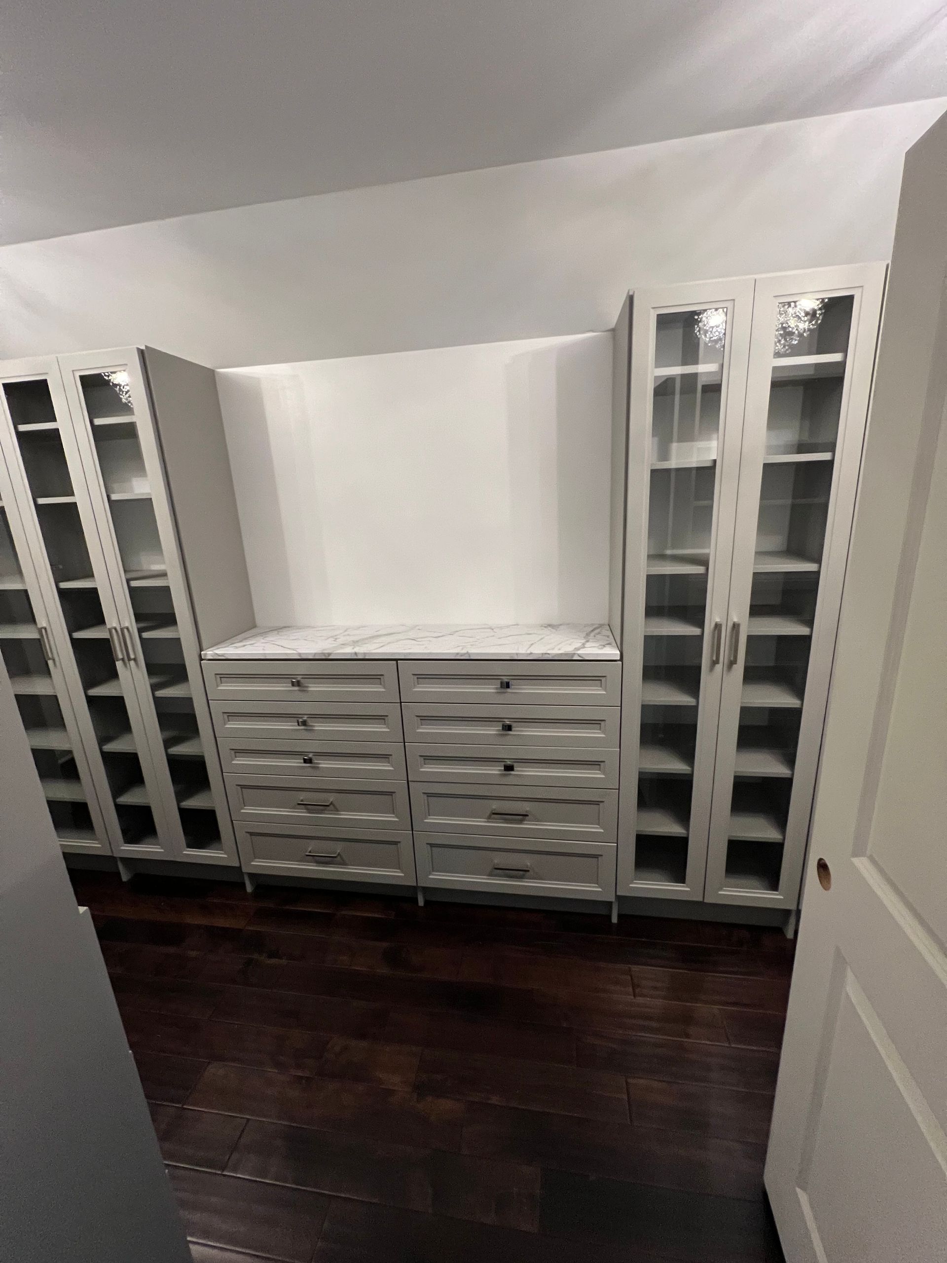 A built-in white walk-in closet featuring glass-door cabinets, drawers with marble-topped counters, and dark wood floors.