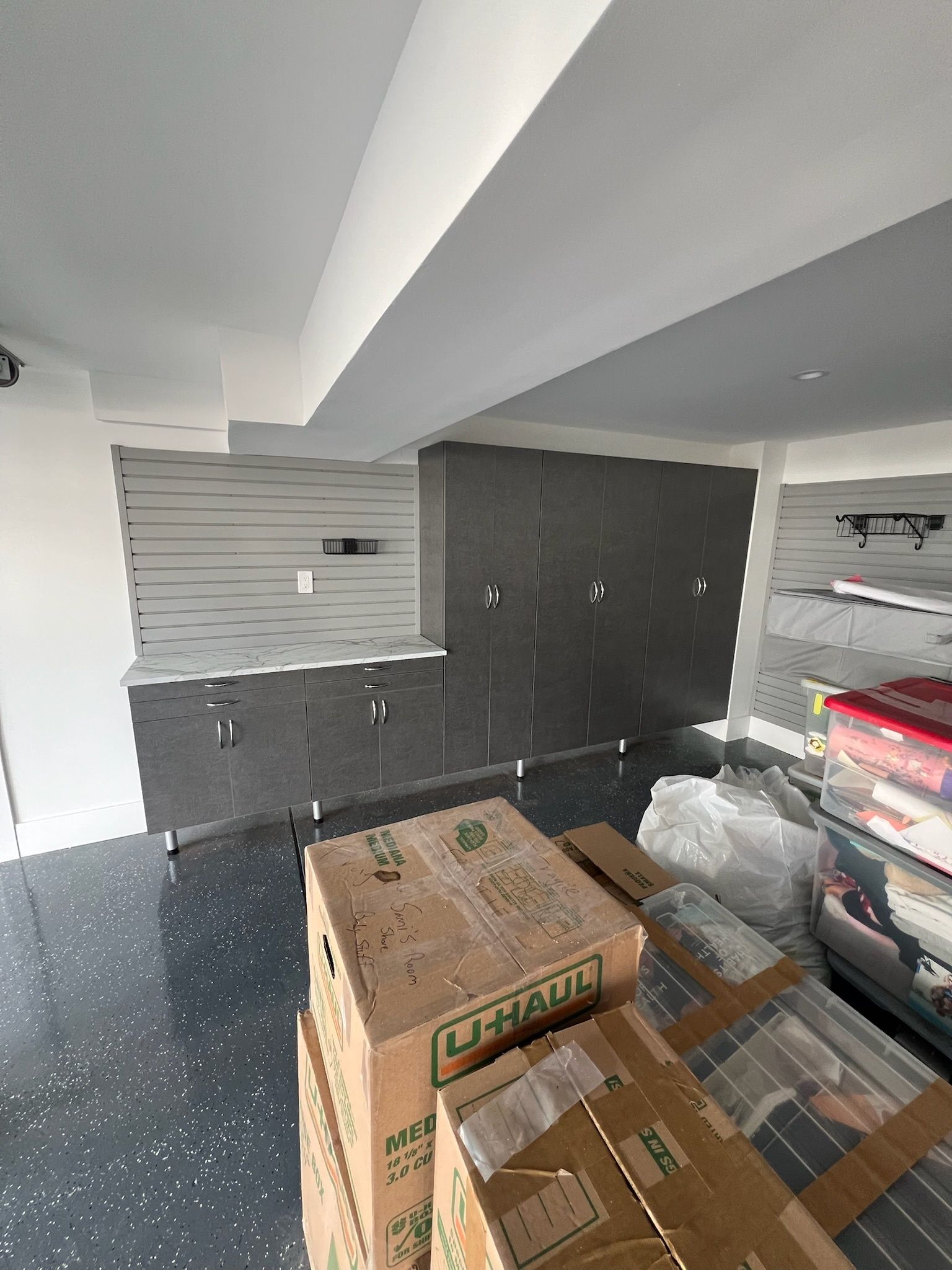A garage interior with gray slat wall panels, dark gray storage cabinets, a speckled floor, and stacked moving boxes.