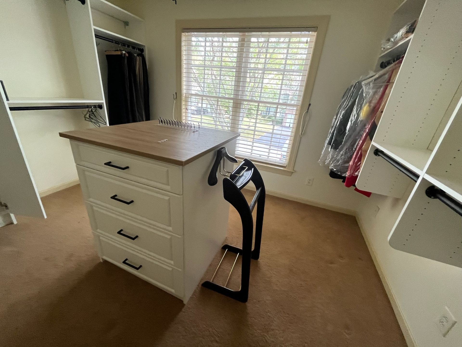 A walk-in closet featuring a central white four-drawer island with a wood top, a valet stand, and clothing racks on walls.