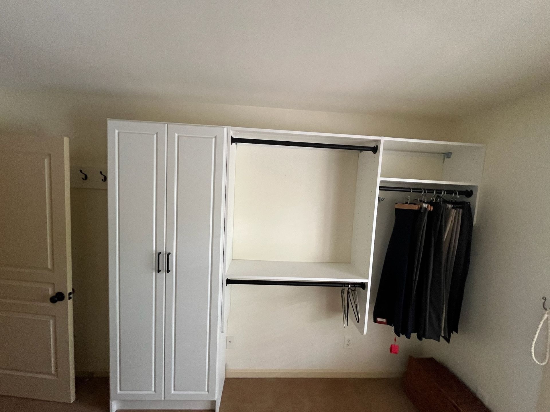 A white modular wardrobe unit with a double-door closet, an open shelf, and a hanging rack against a bedroom wall.