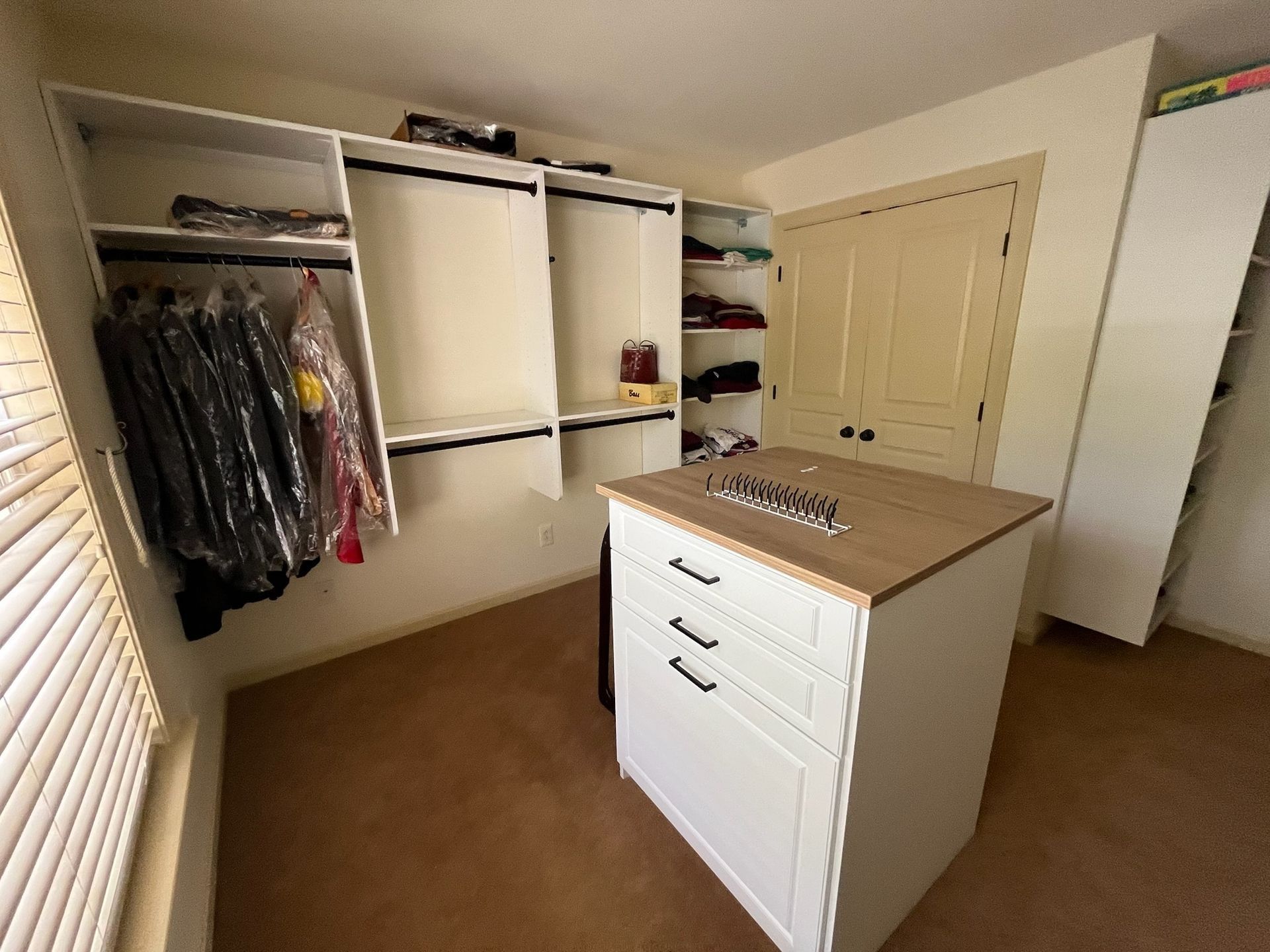 A walk-in closet featuring white built-in shelving, hanging clothes, and a white kitchen island with drawers in the center.