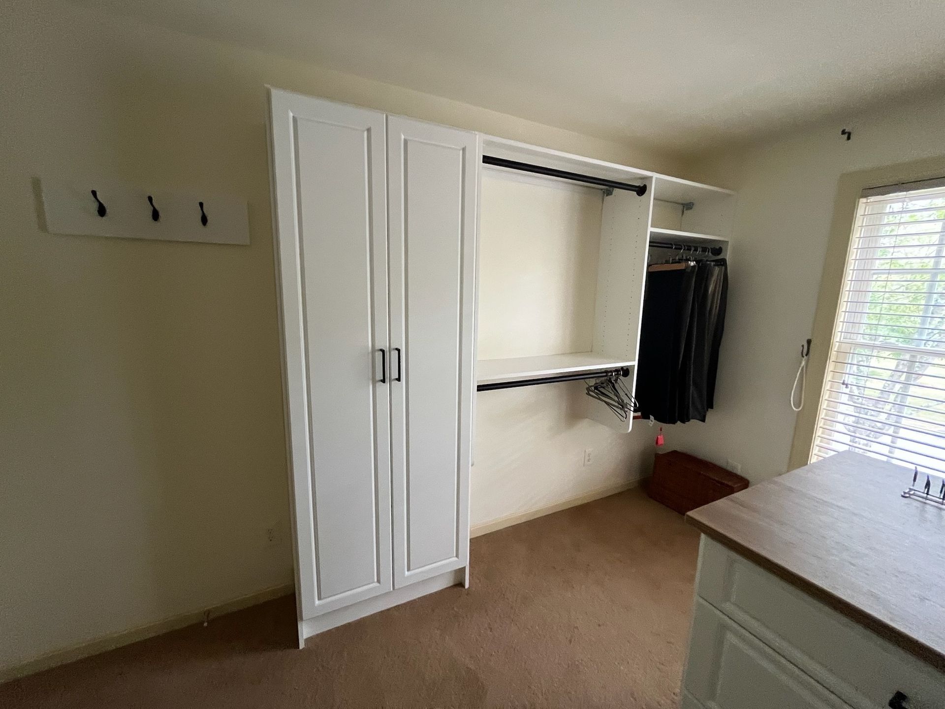A white closet unit with doors and open shelving stands in a carpeted room next to a wall-mounted hook rack and a window.
