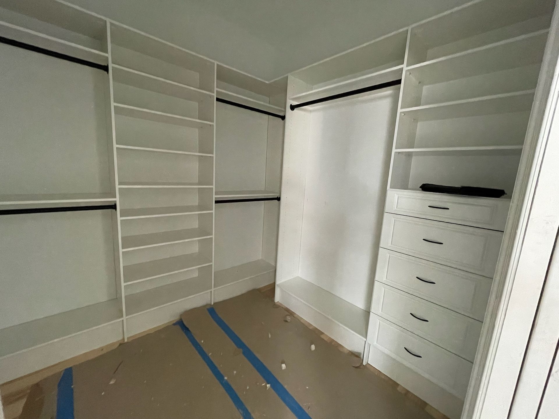 A modern white walk-in closet with shelves, hanging rods, and a stack of five drawers on a floor covered in protective film.