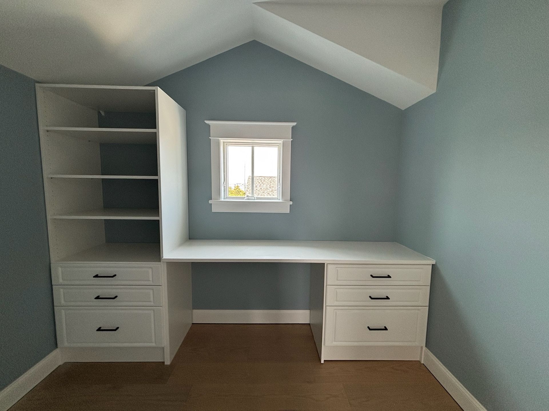 A white built-in desk and shelving unit with drawers, set against a blue wall with a centered window under a vaulted ceiling.