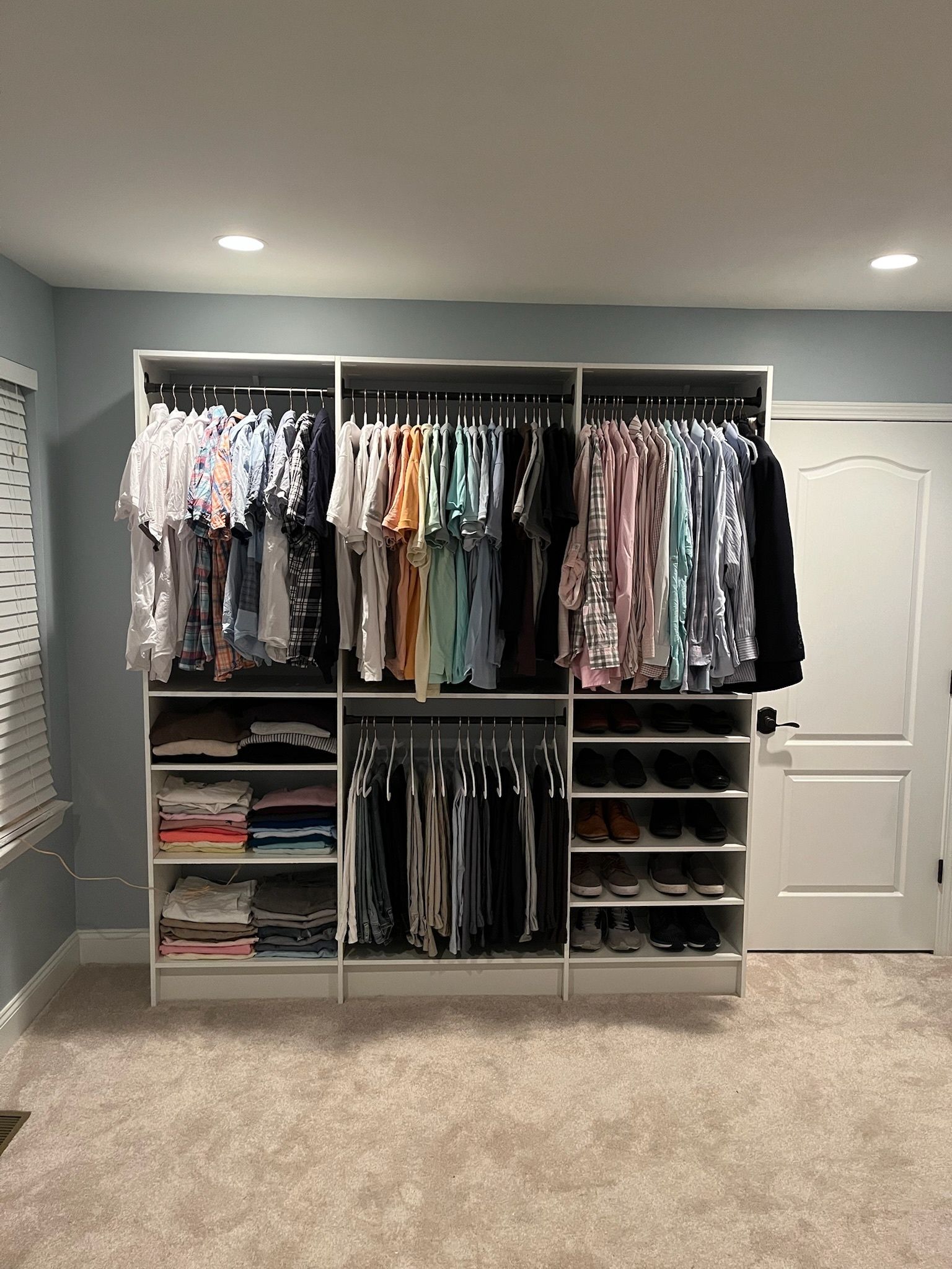A large, white, built-in closet unit against a blue wall, filled with organized hanging clothes and folded stacks.