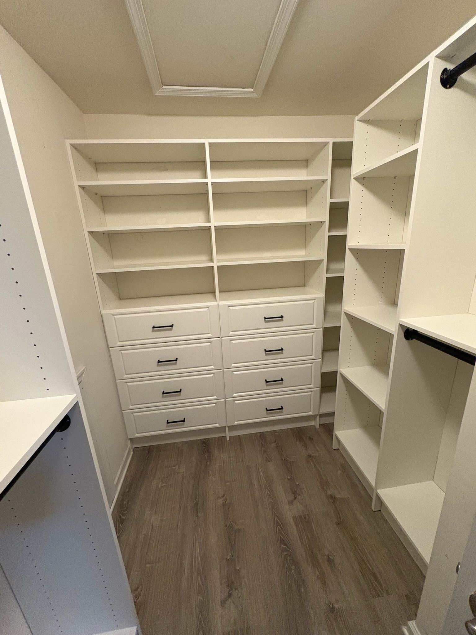 A walk-in closet featuring white wooden shelving units, two columns of drawers, and gray wood-look plank flooring.