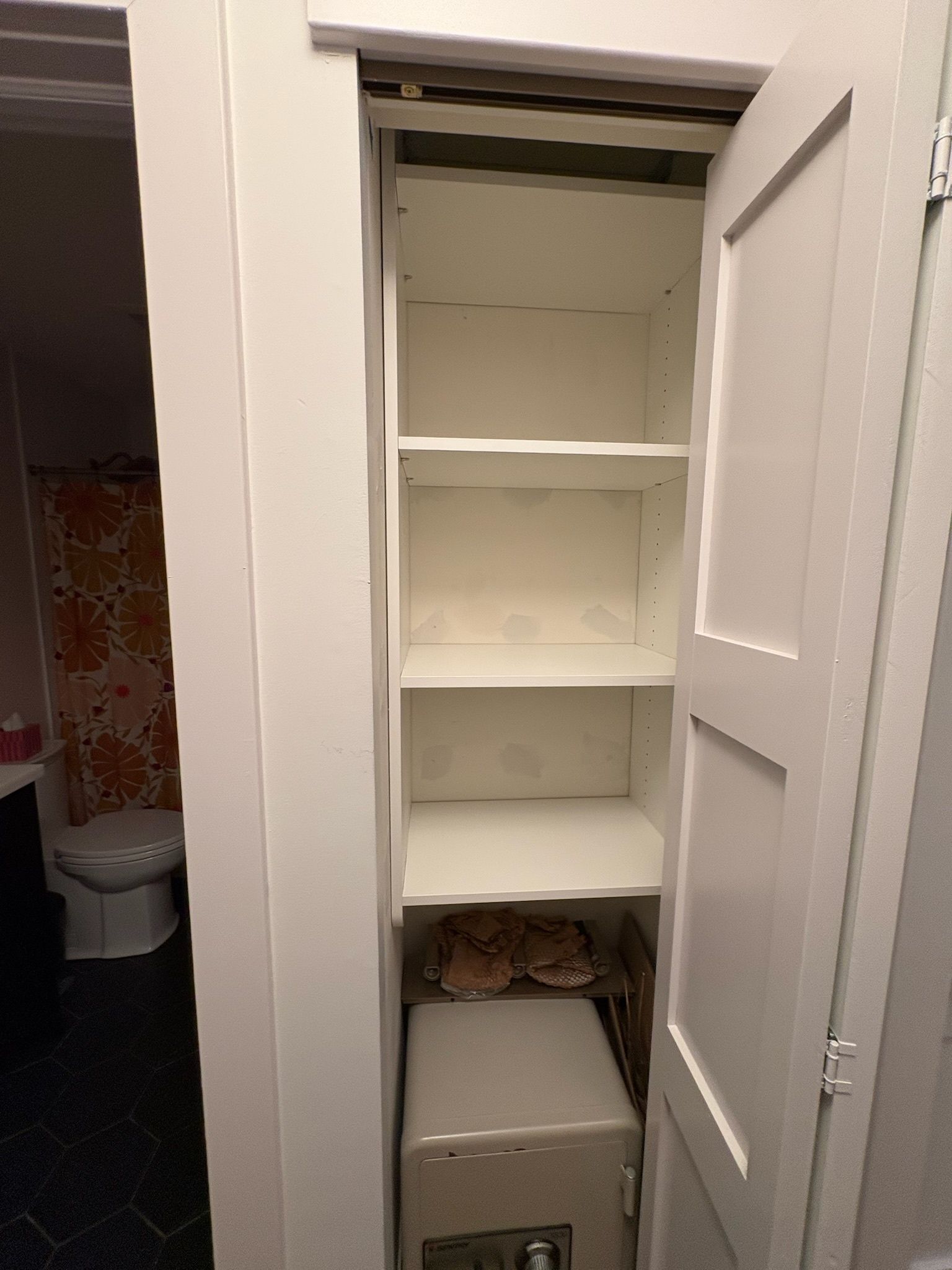 An open white closet with four shelves, featuring a light-colored safe on the bottom shelf, located next to a bathroom.