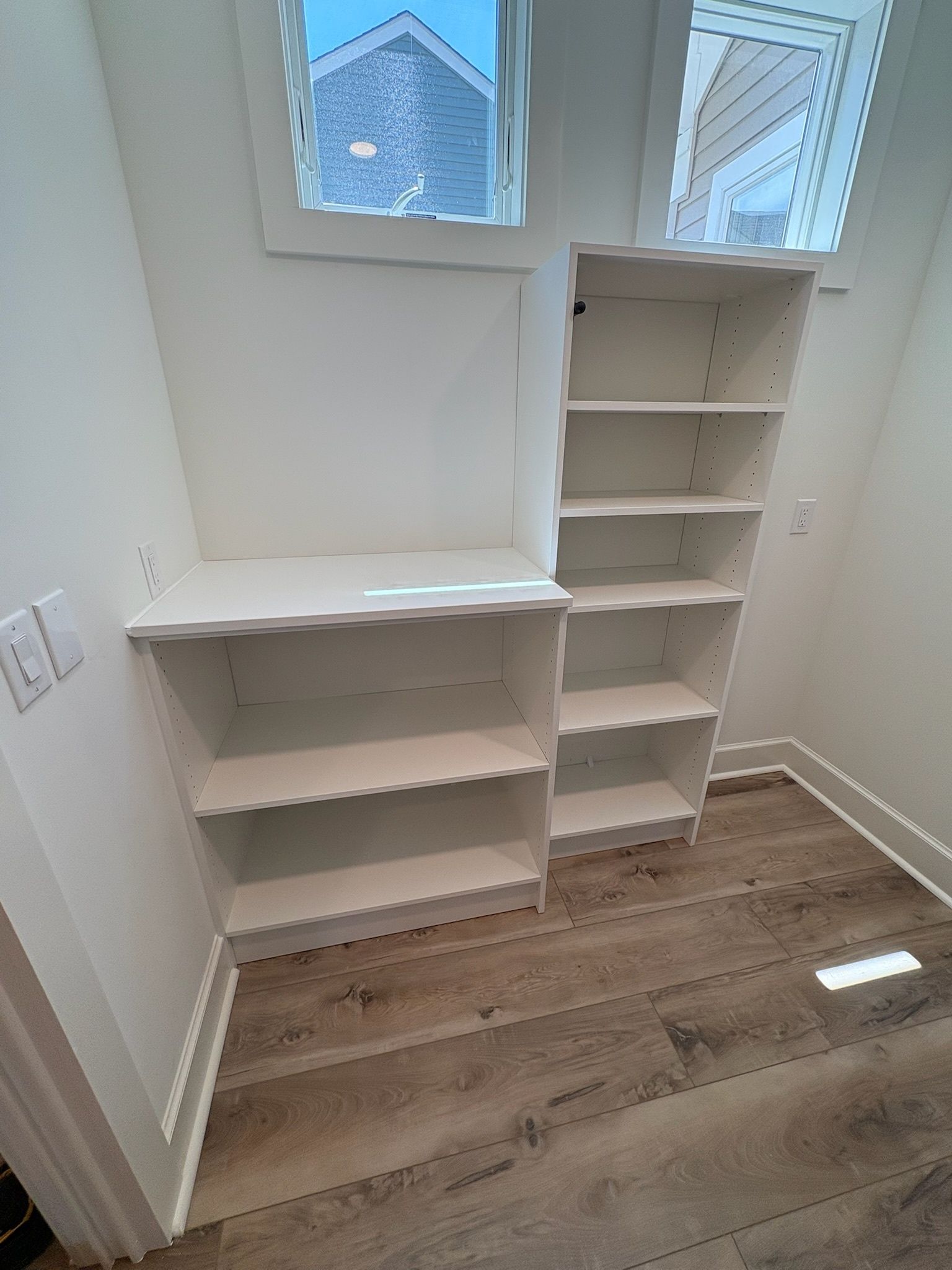 White built-in shelving with a low desk counter and a tall cabinet against a white wall with wood-look flooring.